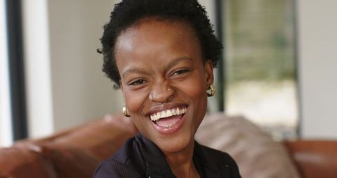 African american woman laughing on leather sofa in cozy living room, candid lifestyle