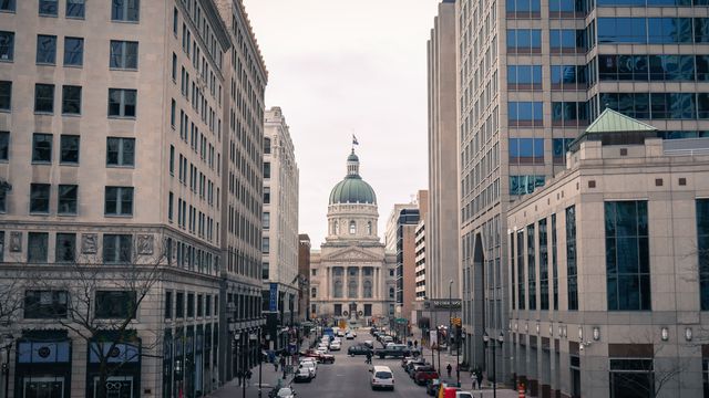 The image shows a vibrant downtown street with a prominent historic government building at the center. Flanked by modern office buildings, the wide street is lined with parked cars and moderate traffic. Ideal for use in articles or projects related to urban development, architecture, government, and city life.