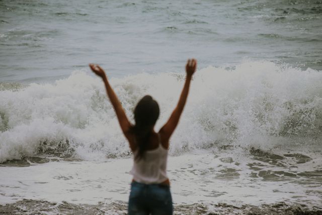 Woman standing on seashore with arms raised facing crashing waves. Perfect for depicting freedom, enjoying nature, summer vacations, coastal activities, or relaxation in natural scenery.