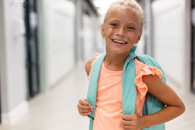 Portrait of cheerful caucasian elementary schoolgirl with backpack standing in school corridor. unaltered, education, childhood, happiness and school concept.