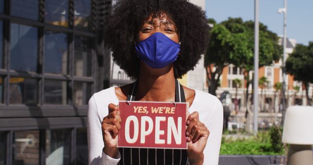 African american female cafe worker wearing face mask showing were open signage. small independent business during coronavirus covid 19 pandemic.