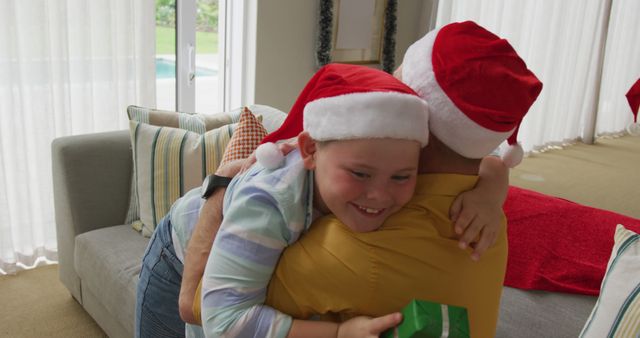 Father and son wearing Santa hats hugging in living room. Son holds a small wrapped gift. Perfect for promotions and advertisements related to Christmas celebrations, family bonding, holiday greetings, or marketing materials highlighting festive joy and family togetherness.