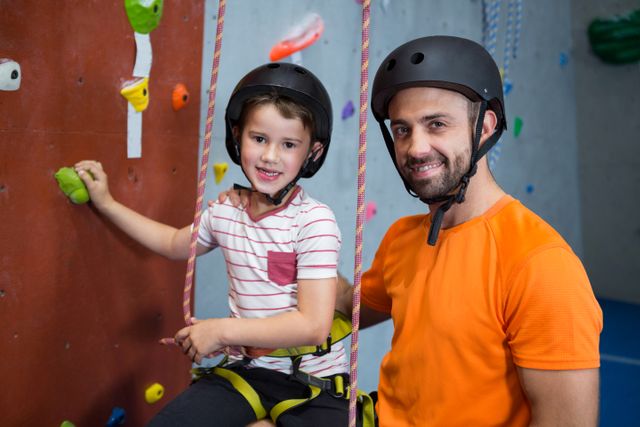 Trainer assisting boy in rock climbing at fitness studio. Both wearing safety gear including helmets and harnesses. Ideal for use in articles about physical fitness, indoor sports, training, mentorship, and family bonding activities.