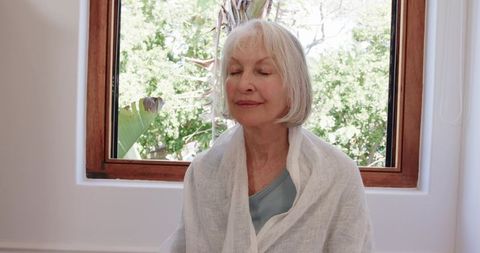 Senior woman meditating near sunlit window in white room