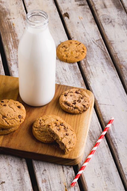 This image shows a high angle view of a milk bottle and chocolate chip cookies on a rustic wooden table. A red striped straw is placed beside the cookies. Ideal for use in food blogs, recipe websites, advertisements for dairy products, or healthy eating promotions. The rustic setting adds a homely and comforting feel, perfect for marketing homemade or artisanal products.