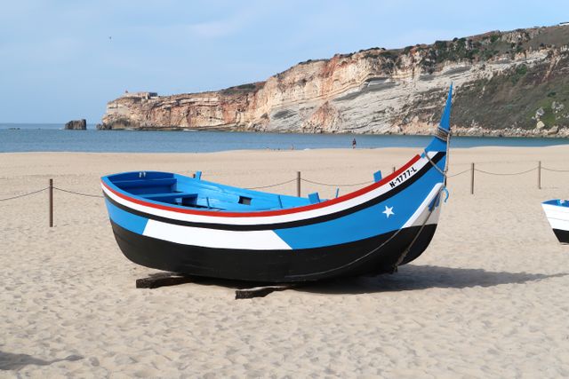 A traditional, colorful fishing boat rests on the sandy beach with rocky cliffs in the background. This scene reflects peaceful coastal beauty, making it ideal for travel brochures, tourism promotions, or marine-themed publications.