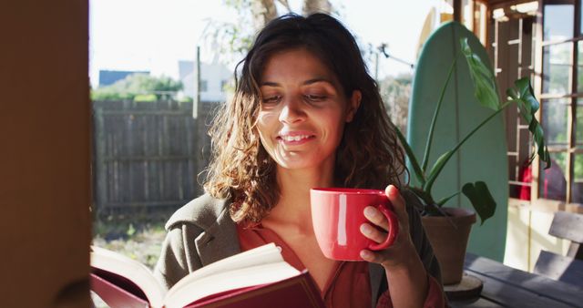 Happy biracial woman sitting drinking tea and reading book in sunny cottage kitchen. simple living in off the grid rural home.
