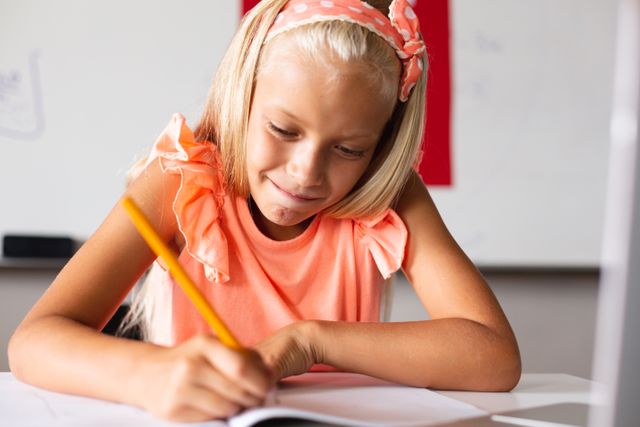 Smiling caucasian elementary schoolgirl writing on book while sitting at desk in classroom. unaltered, education, learning, studying and school concept.