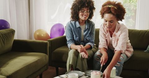 African American friends celebrating at home cutting cake on green sofa with balloons
