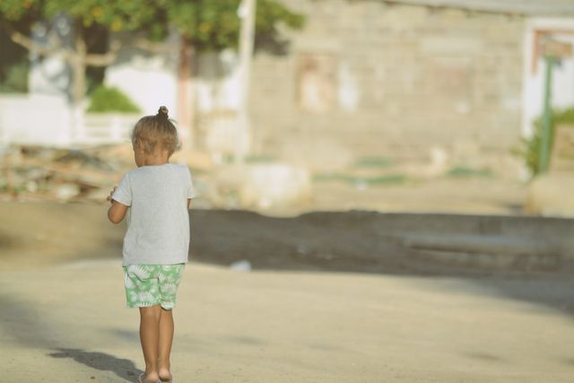 Little girl is walking away in a sunlit village. She is clad in casual clothing, wearing shorts and a t-shirt, with her hair up in a bun. Background includes a blurry rural setting with houses and trees. Good for concepts of childhood, innocence, rural life, simplicity, and exploration. Suitable for use in articles, blogs, or advertisements related to countryside living, solo outdoor activities, and exploration.