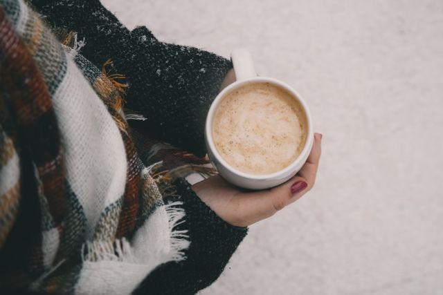 Person holding cup of coffee while wrapped in a scarf on a snowy day. Perfect for themes related to winter, coziness, relaxation, and outdoor activities. Ideal for use in articles, blogs, and advertisements about coffee or seasonal lifestyles.