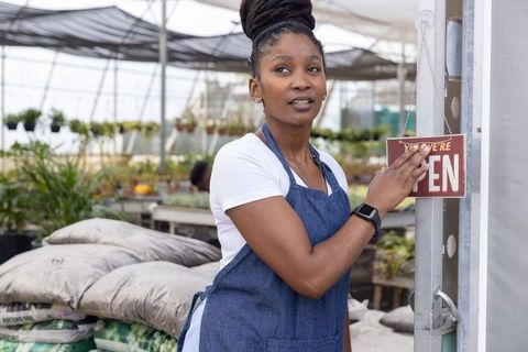 African american woman placing open sign at greenhouse nursery, small business owner in apron
