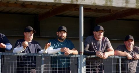 Baseball teammates leaning on dugout fence watching game in sunny ballpark