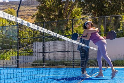 Hugging over padel net on sunlit outdoor court, holding paddles, celebrating friendship