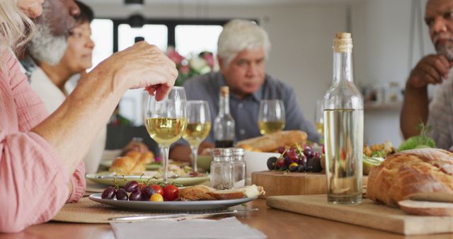 This image depicts a group of senior friends enjoying lunch together at a dining table. The table is set with wine, bread, cheese, and a plate of grapes, indicating a casual and relaxed meal. One person is reaching to serve themselves, while the others chat and enjoy each other's company. This image can be used for articles about socializing in retirement, dining with friends, or healthy eating habits among older adults.