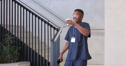 African american clinician in navy scrubs holding coffee and smartphone on staircase