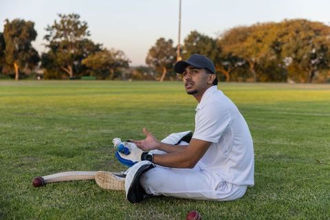 Indian cricketer sitting on grass wearing pads and cap with bat and red ball at sunset