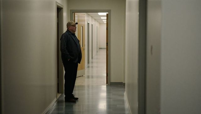 Older man waiting in quiet institutional corridor, hands in pockets, contemplative mood