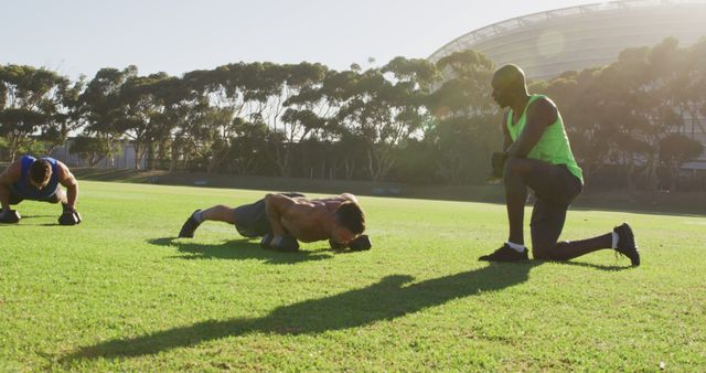 Two men performing push-ups on grassy field with personal trainer assisting on a sunny day. Ideal for content related to fitness and health, outdoor exercise routines, personal training services, and active lifestyles.
