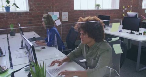 Typing curly-haired professional working at modern coworking desk behind clear plexiglass partition
