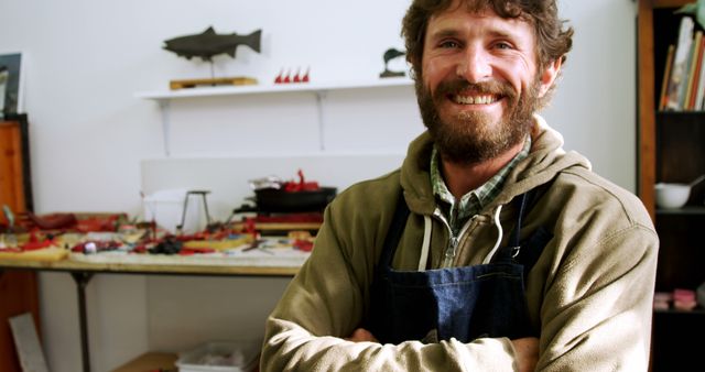 A smiling Caucasian man in a workshop setting, wearing a jacket and apron, suggests he might be a craftsman or artist, with copy space. His cheerful expression and casual attire convey a sense of satisfaction and creativity in his work environment.