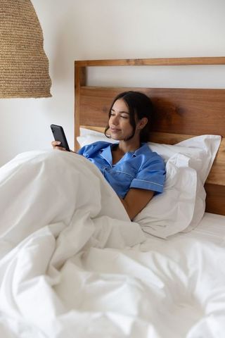 Woman reclining in bed wearing blue pajamas scrolling smartphone in minimalist bedroom