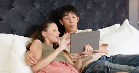 Young couple relaxing in bed sharing tablet, laughing and browsing together