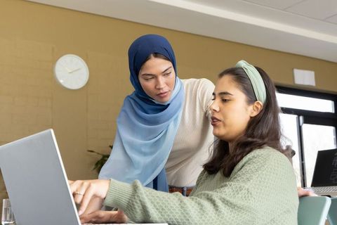 Diverse female colleagues collaborating and reviewing work over laptop in modern office