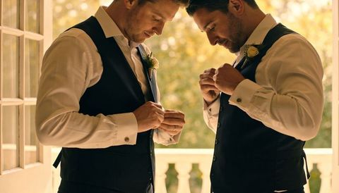 Groomsmen fastening cufflinks and adjusting waistcoats on sunlit balcony before ceremony