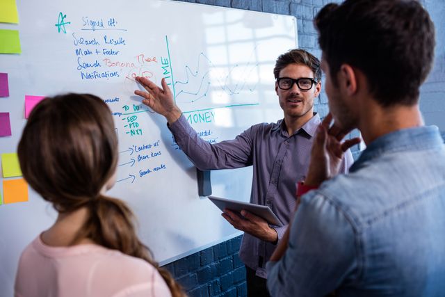 Coworkers are collaborating in a modern office environment, discussing ideas and strategies in front of a whiteboard filled with notes and graphs. This image can be used to illustrate teamwork, business meetings, project planning, and professional collaboration in corporate settings.