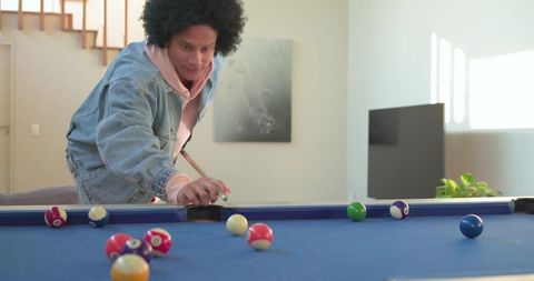 Mid-adult man leaning over pool table aiming cue wearing denim jacket and pink hoodie