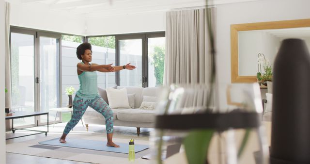 Afro woman stretches in living room practicing yoga on mat, surrounded by modern furniture. Suitable for content promoting home workouts, wellness lifestyles, physical health, indoor sports, and mental relaxation.