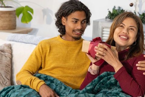Couple cuddling on sofa holding wrapped gift, wearing knit sweaters under teal blanket