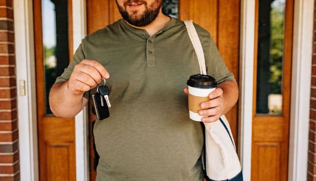 Bearded man handing car keys at front door holding coffee cup and canvas bag
