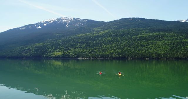 Two individuals are kayaking on a tranquil lake with a backdrop of lush green hills and snow-capped mountains. Engaging in water sports amidst such serene natural beauty offers a perfect opportunity for relaxation and adventure.