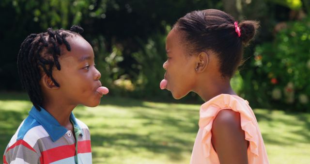 Two young children playfully sticking their tongues out at each other in a garden, capturing a moment of childhood fun. The image can be used for themes revolving around play, youth, and outdoor activities, making it ideal for marketing campaigns, educational materials, and articles about child development.