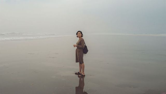 Solo woman standing on reflective wet sand looking back on misty overcast beach