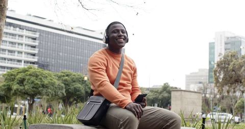 African American man smiling listening to music on headphones in urban park holding smartphone