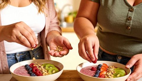 Two women decorating smoothie bowls with berries, kiwi and granola