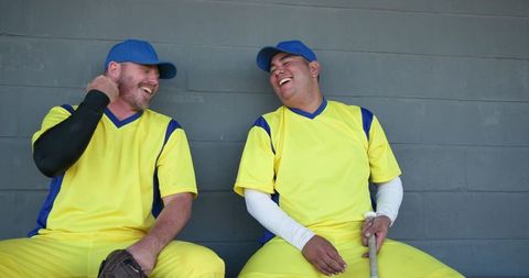 Baseball teammates laughing on dugout bench wearing yellow jerseys and blue caps