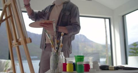 Indian male painter selecting brush at easel in bright lakeside studio with palette