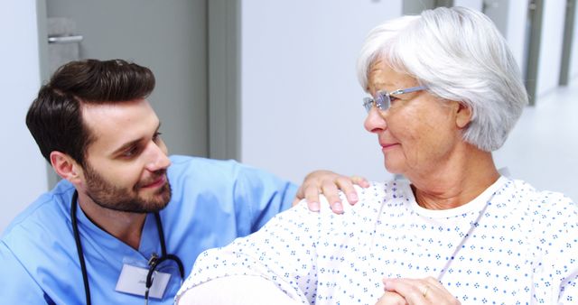 Male nurse wearing blue scrubs comforting elderly female patient with gray hair in hospital corridor. The nurse's hand rests gently on the patient's shoulder, showing support and compassion. Perfect for healthcare, elderly care, and medical related themes. Useful for articles, blogs, and advertisements focusing on compassionate care in medical settings.