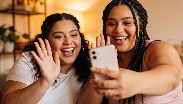 Black women friends smiling and waving during video call on cozy living room couch
