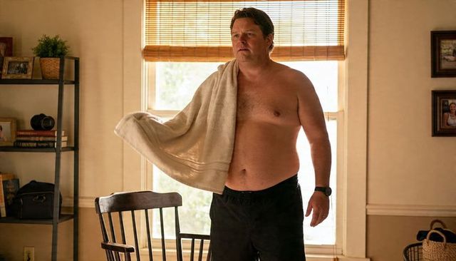 Shirtless man standing with towel over shoulder in black shorts and wristwatch in sunlit room