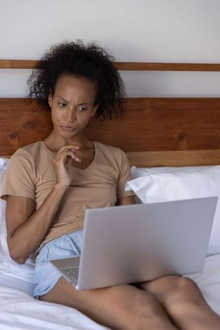 African american woman working on laptop in bed looking thoughtful and puzzled at screen