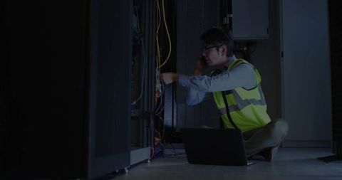 Technician Working in Dimly Lit Server Room with Cables and Laptop