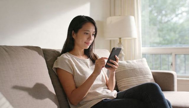 Asian woman relaxing on sofa checking smartphone in sunlit living room for remote tasks