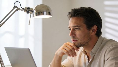 Focused man working on laptop in minimalist home office with chrome desk lamp