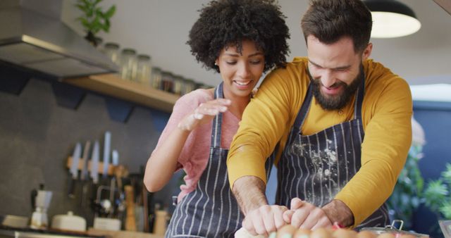 Image of happy diverse couple in aprons baking in kitchen, kneading dough, with copy space. Happiness, communication, inclusivity, free time, togetherness and domestic life.