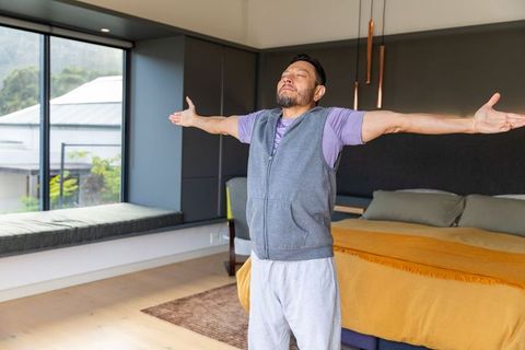 Mature asian man stretching and breathing deeply in modern minimalist bedroom by window